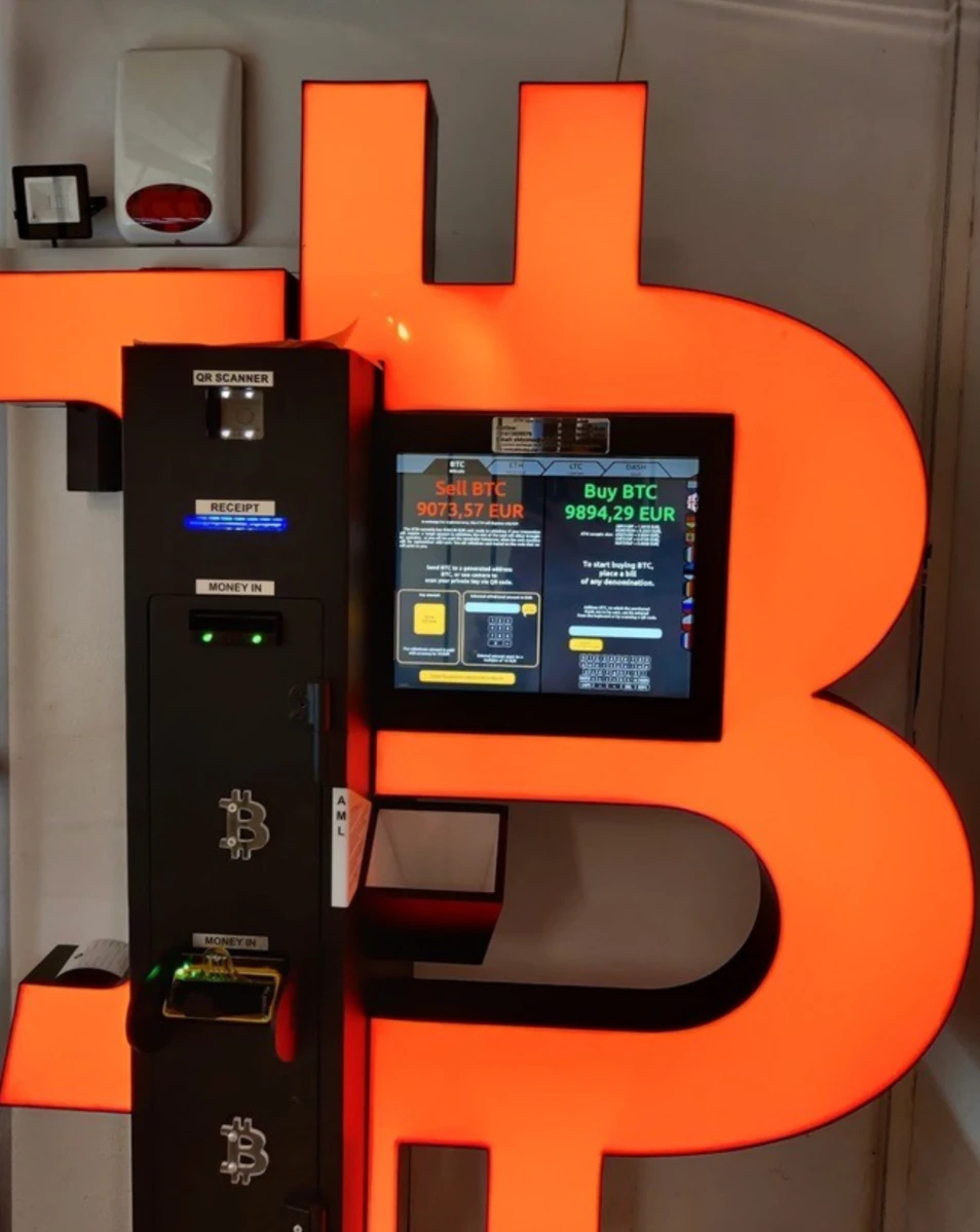 Distributeur automatique de Bitcoin avec &eacute;cran tactile affichant les taux d&rsquo;achat et de vente en euros, install&eacute; devant un grand logo lumineux orange en forme de symbole BTC