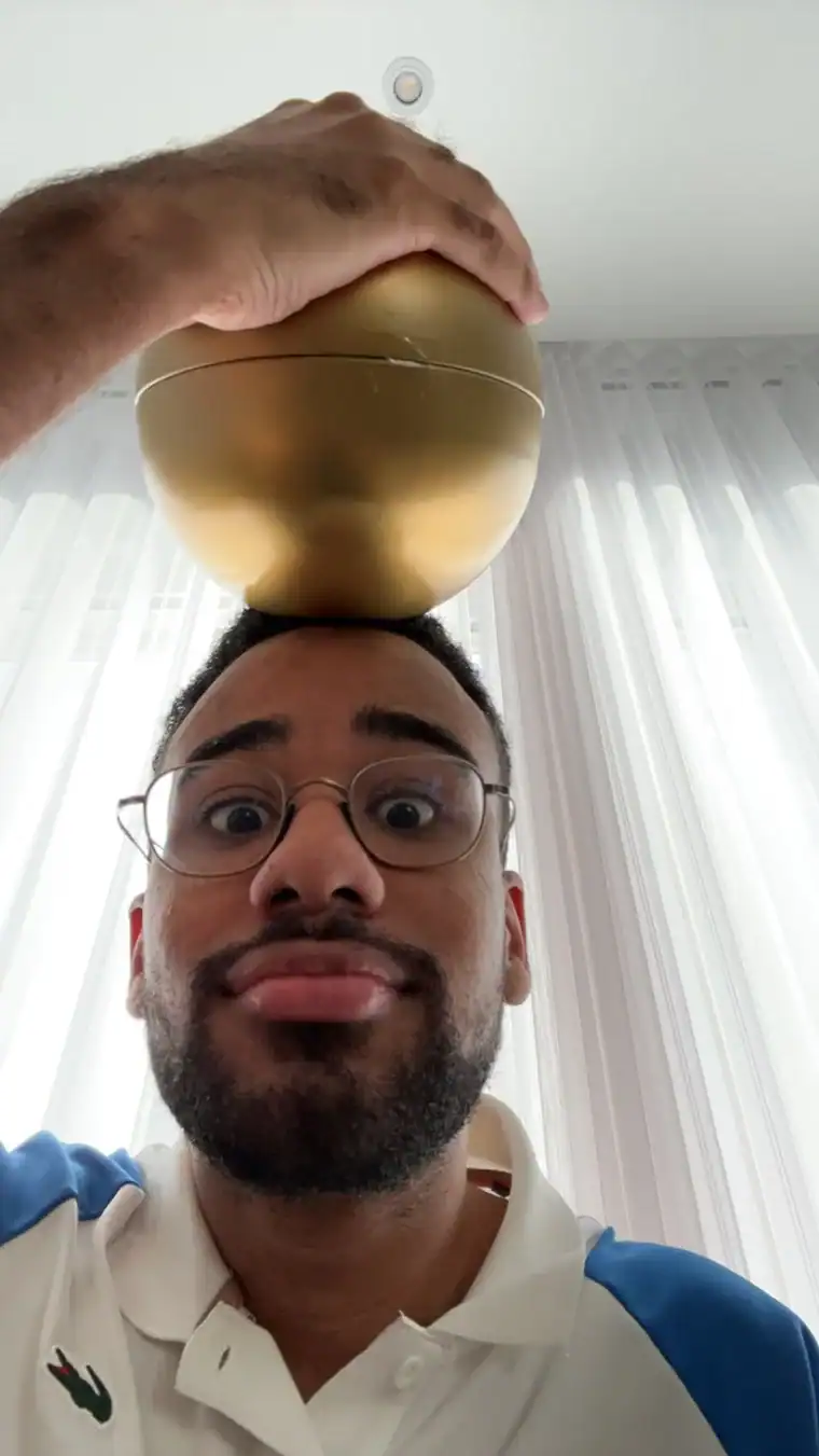 Photo de NeverSayYes avec un bol doré en équilibre sur la tête et des lunettes rondes, polo blanc et bleu.